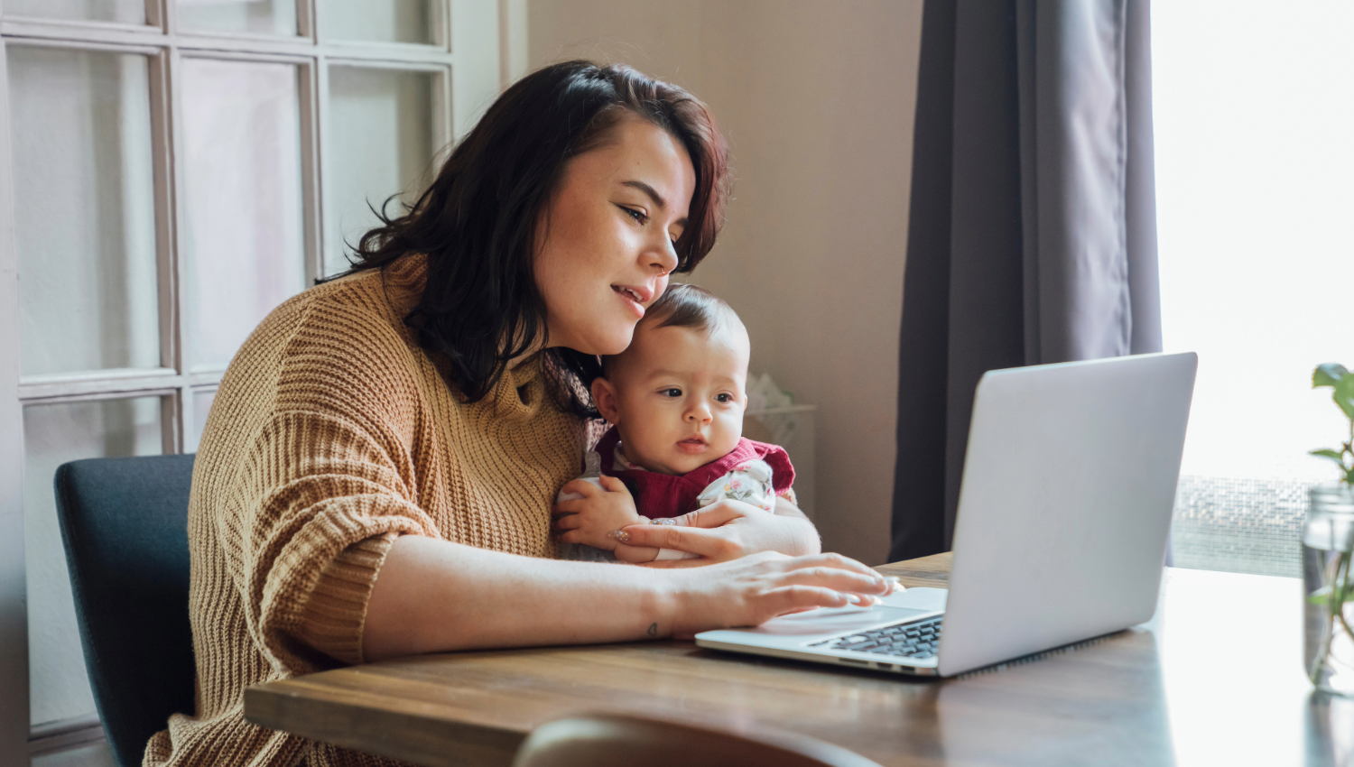 Mom holding baby and working from home on laptop at dining room table